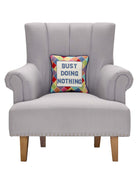 Busy Doing Nothing Embroidered Needlepoint Throw Pillow on an armchair, showing the throw pillow at room scale, 12x12 inch
